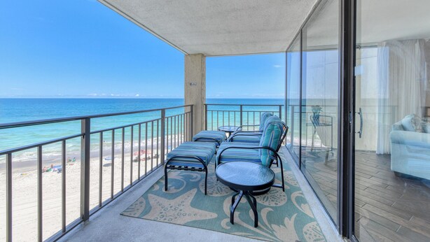 A balcony with a view of the ocean, furnished with chairs and a table, and a glass door leading to a living area.