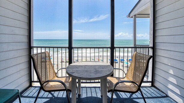 Un balcon avec une table et deux chaises, donnant sur une plage parsemée de parasols et fréquentée par des gens.