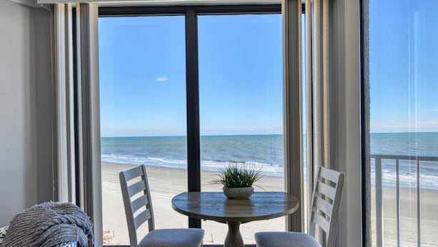A room with a large window overlooking a beach, two chairs, a small round table with a plant, and a cozy seating area with a blanket. - One Ocean Place 403 - Fantastic two-bedroom, two-bath oceanfront condo in GCB (Garden City Beach)