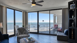 A spacious living room with a large window overlooking a beach, a ceiling fan, and a view of the ocean. - One Ocean Place 403 - Fantastic two-bedroom, two-bath oceanfront condo in GCB (Garden City Beach)
