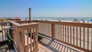 Une terrasse en bois avec une rambarde en métal, donnant sur une plage où l’on aperçoit des parasols et des gens.