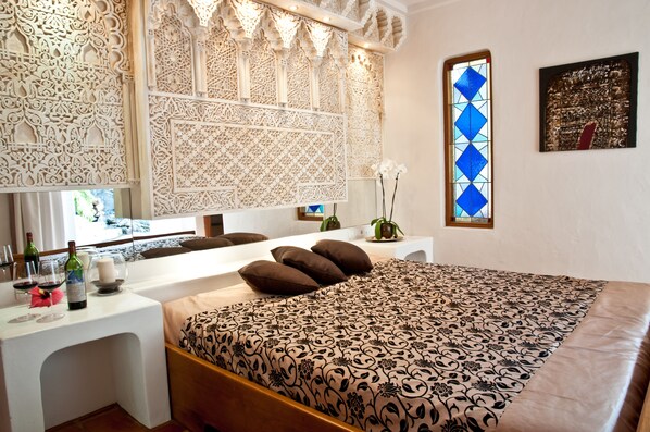 A bedroom with a patterned bedspread, a white bedside table with wine bottles, and a decorative wall panel.