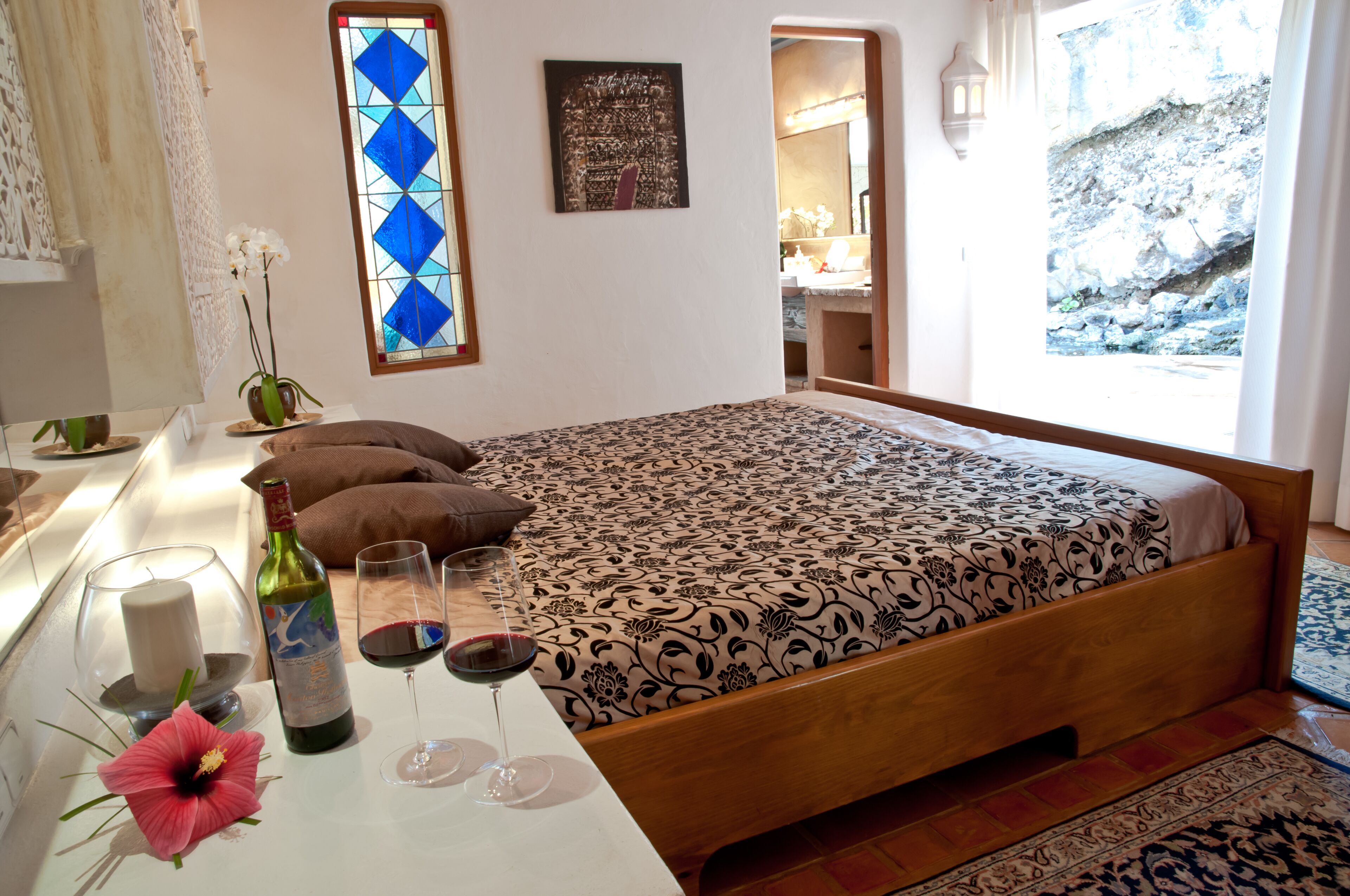 A bedroom with a bed, a nightstand, a vase with flowers, a bottle of wine, and two wine glasses.