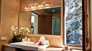 Ein Badezimmer mit einem großen Spiegel, einem Waschbecken, einer Orchidee im Topf und Blick auf einen Wasserfall durch ein Fenster.
