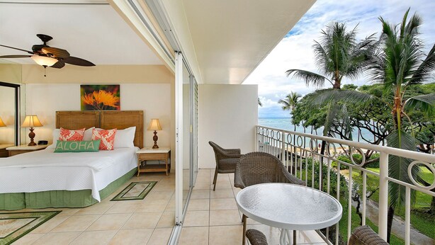 Une chambre d’hôtel avec un lit, un ventilateur de plafond, un balcon avec une table et des chaises, et une vue sur l’océan et les palmiers.