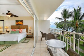 Een hotelkamer met een bed, een plafondventilator, een balkon met een tafel en stoelen, en uitzicht op de oceaan en palmbomen.