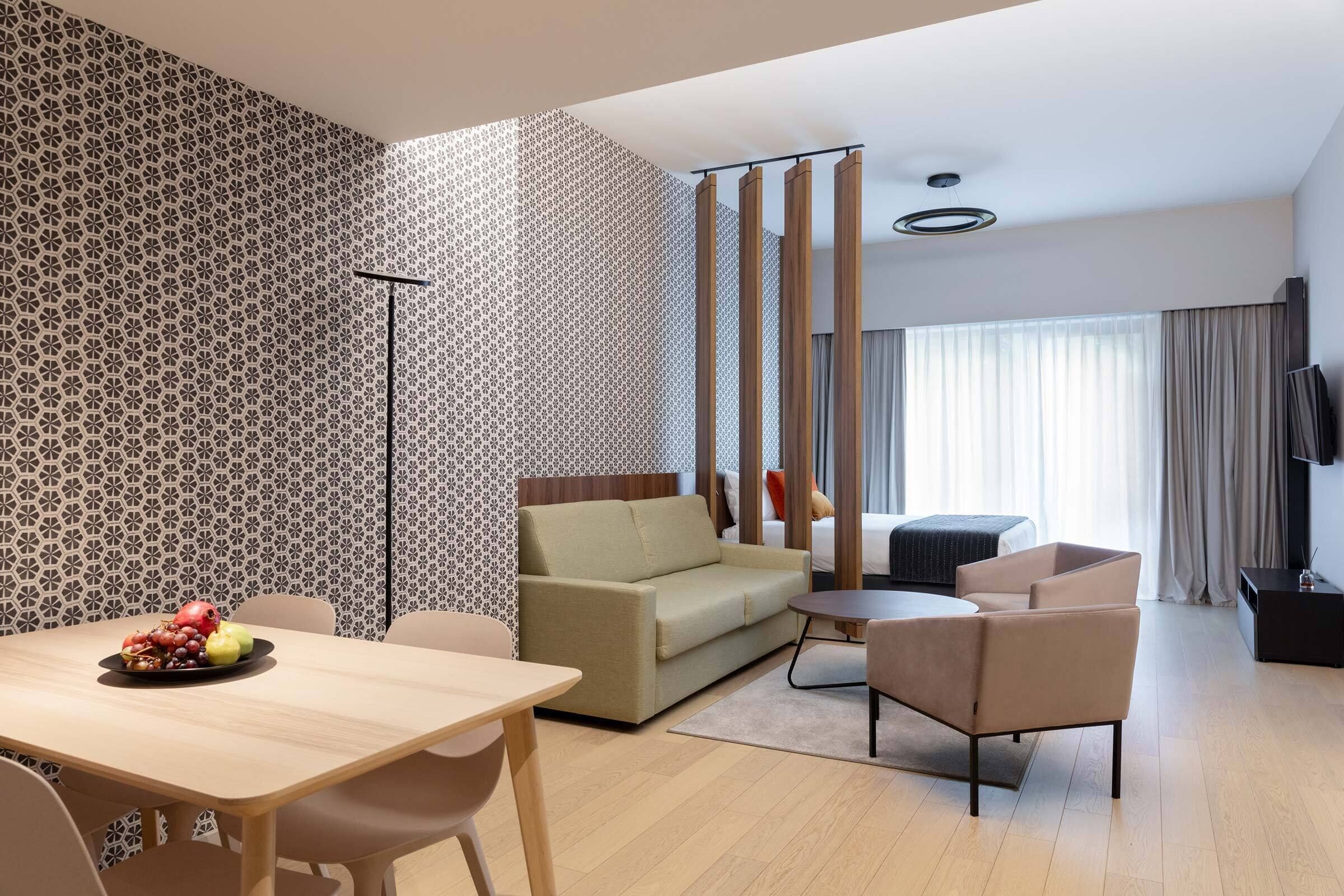 Ein modernes Hotelzimmer mit einer Couch, einem Esstisch mit Obst, einem Fernseher und einem großen Fenster mit Vorhängen.