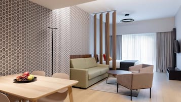 Una moderna camera d'hotel con divano, un tavolo da pranzo con frutta, televisore e un'ampia finestra con tende.