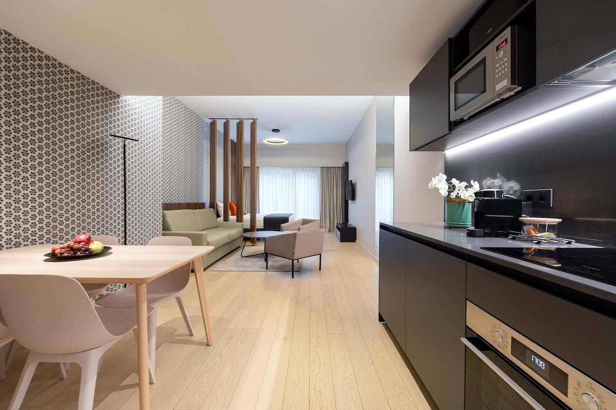 A modern kitchen with a microwave, oven, and a digital clock. There is a dining area with a table and chairs, and a sofa in the background.