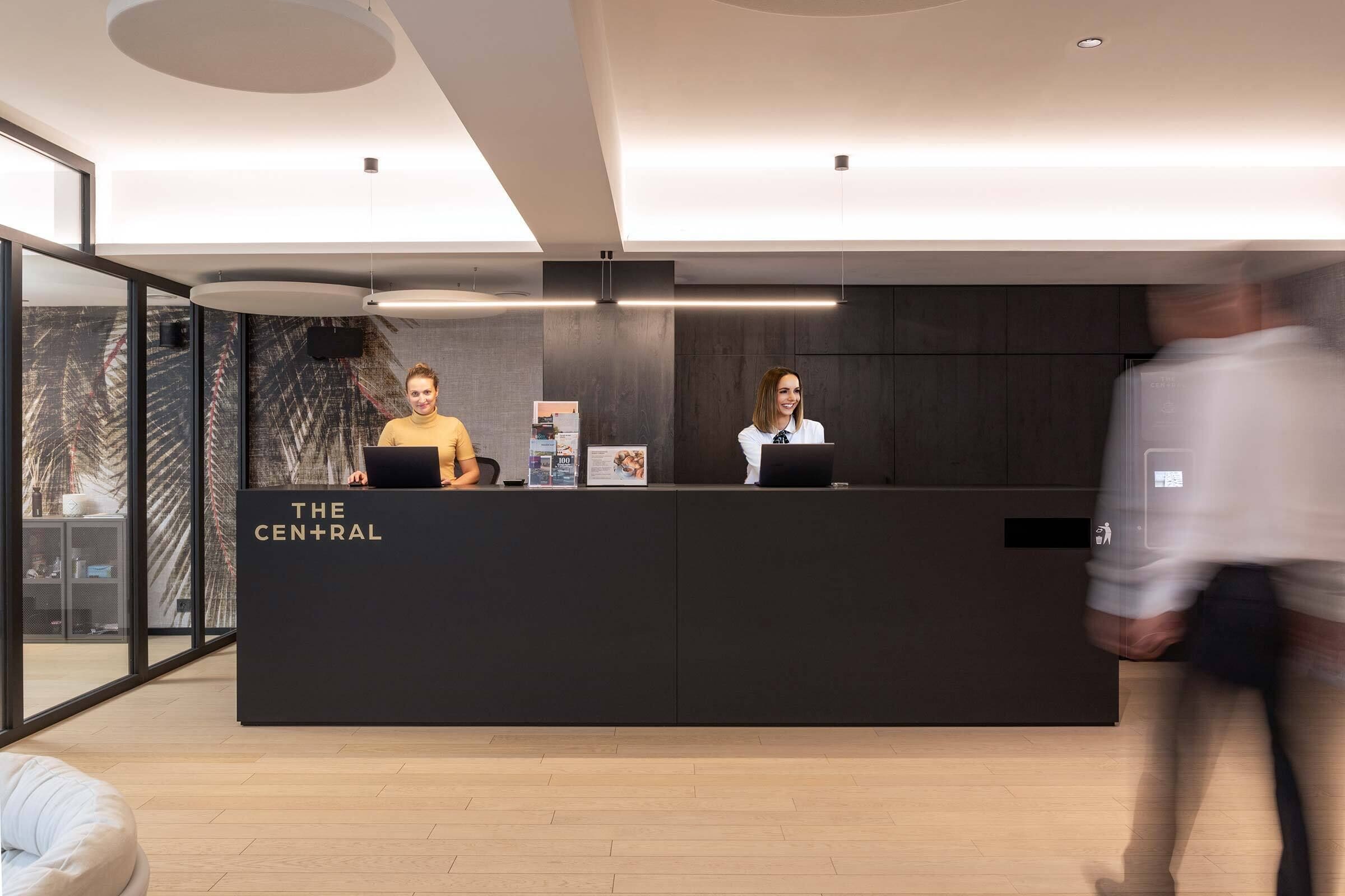 A reception desk with two staff members, a modern interior, and a glass wall.