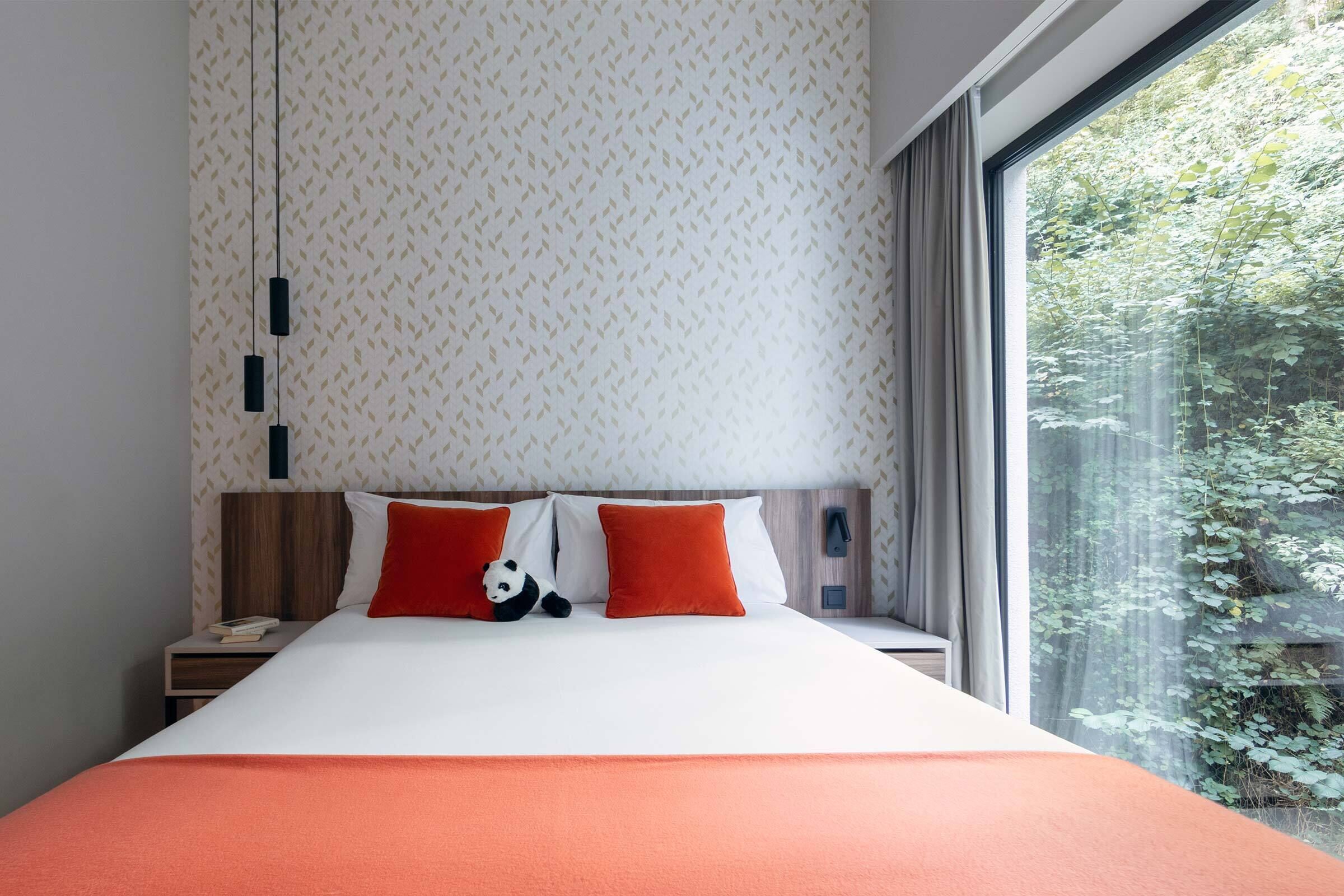 Ein modernes Schlafzimmer mit einem großen Bett, zwei orangefarbenen Kissen, einem Plüschpanda und Blick auf Grün durch ein großes Fenster.