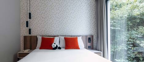 Een moderne slaapkamer met een groot bed, twee oranje kussens, een pluche panda en uitzicht op groen door een groot raam.