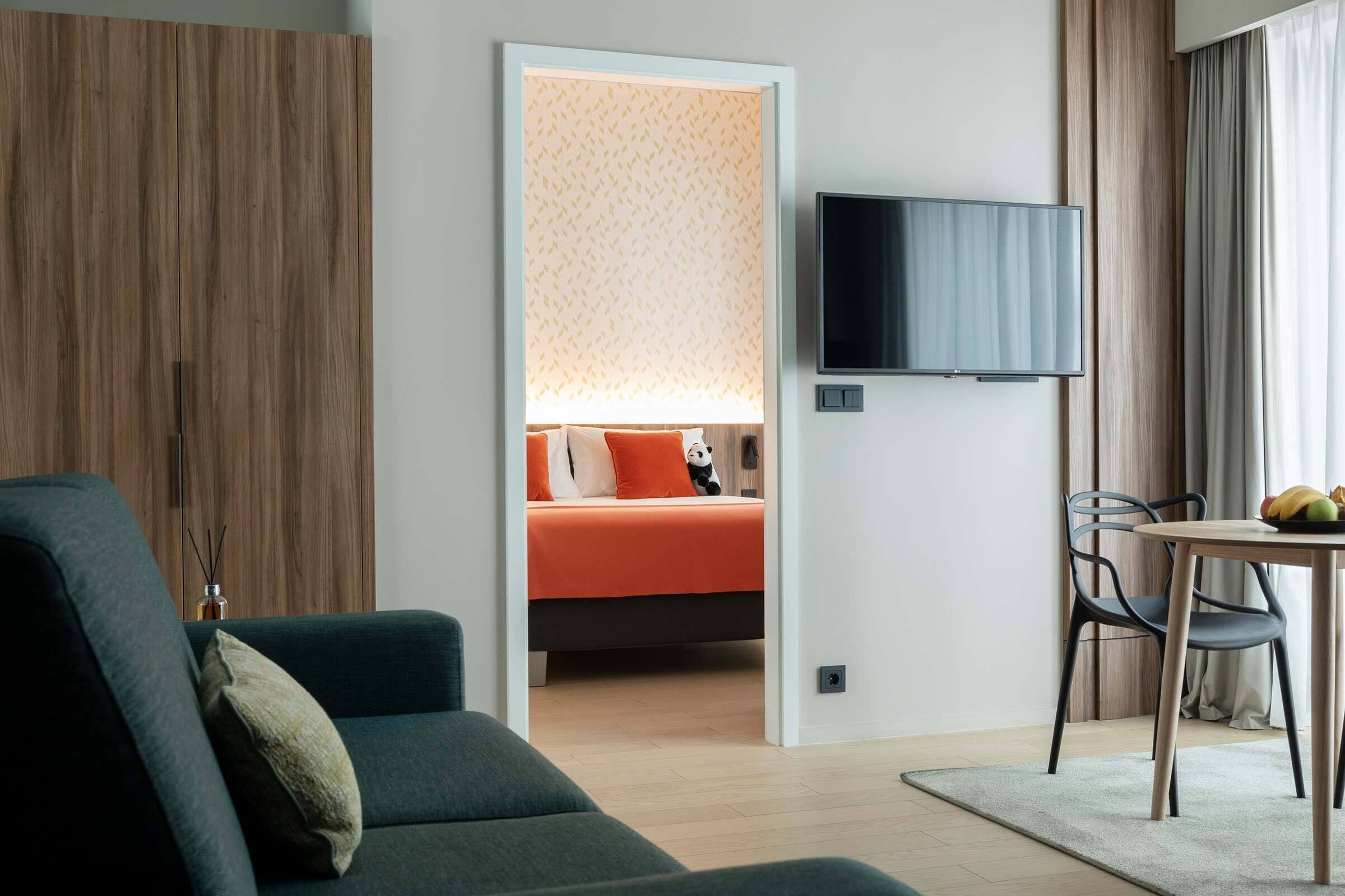 Une chambre d’hôtel moderne avec un canapé, un lit avec une literie orange, un téléviseur fixé au mur et un coin repas comprenant une table et des chaises.