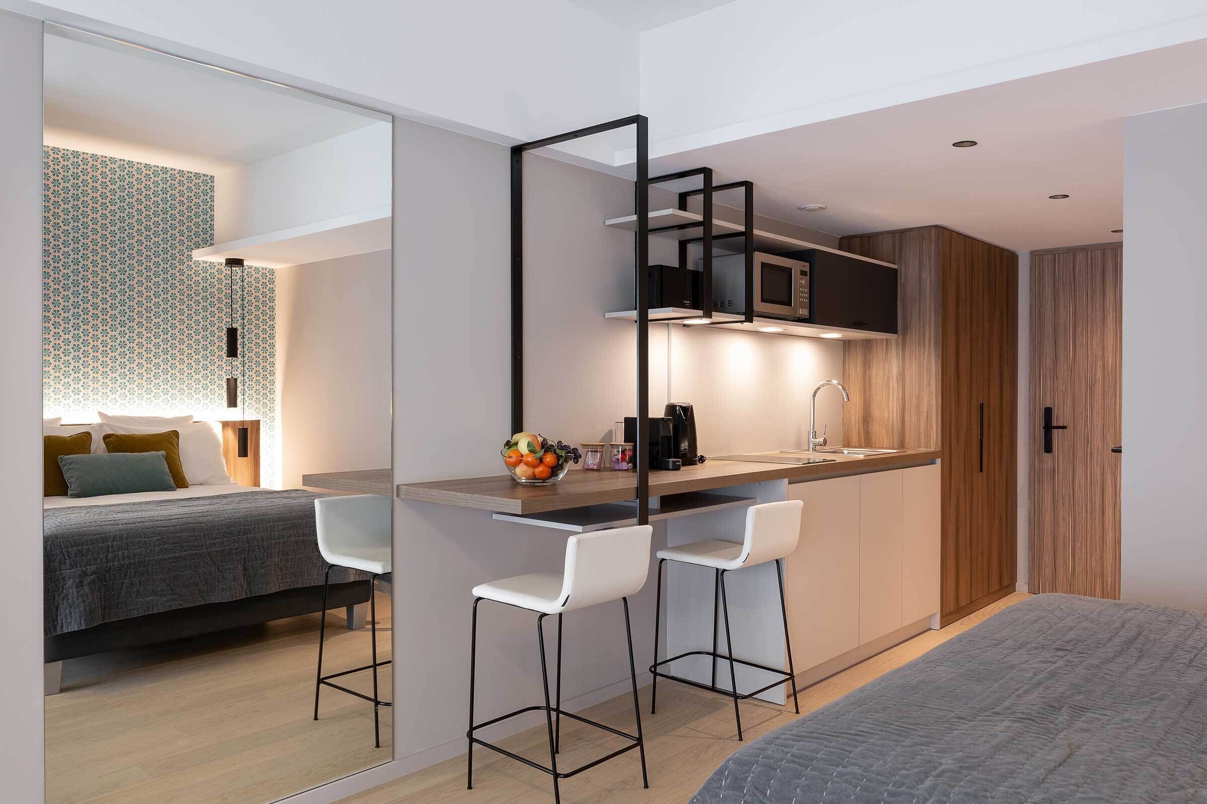 Habitación de hotel moderna y compacta con kitchenette, una cama con cubrecama gris, una silla blanca y un pequeño comedor.