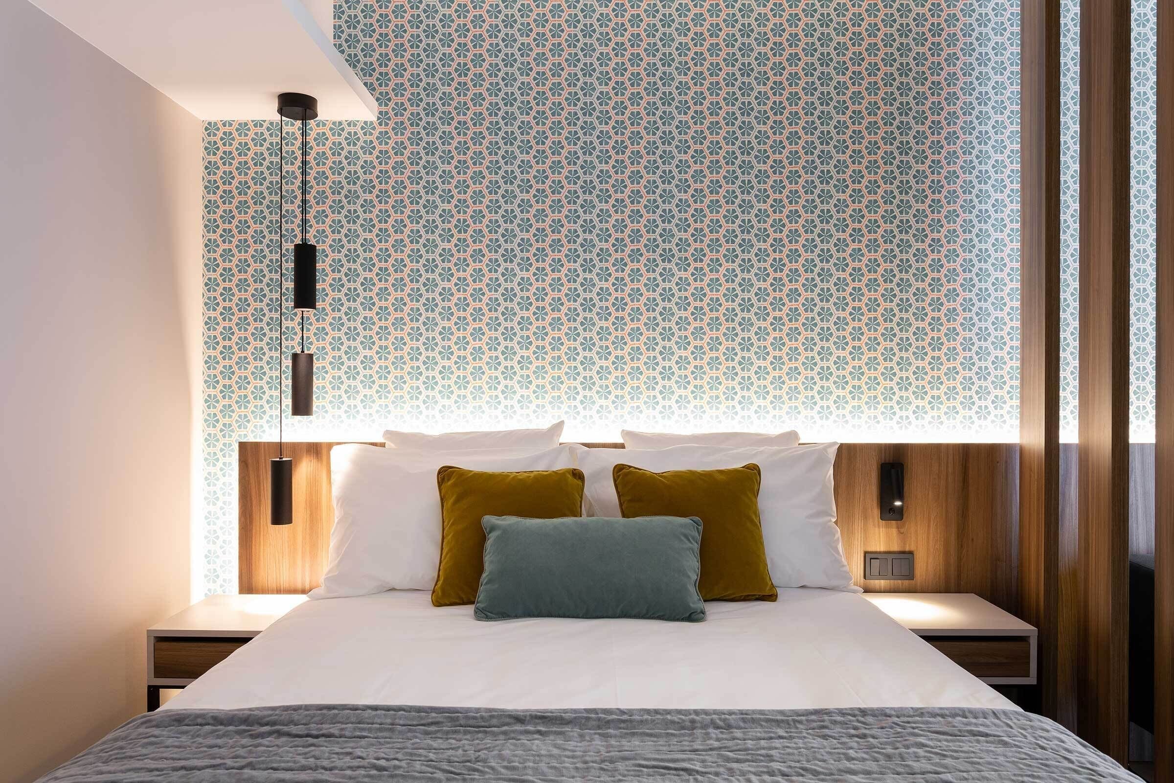 Een moderne hotelkamer met behang met een patroon, een opgemaakt bed met twee kussens, een nachtkastje met een lamp en een houten hoofdbord.
