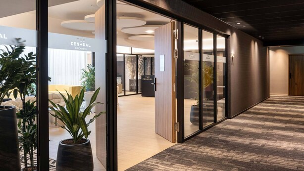Un pasillo moderno de hotel con puertas de cristal, plantas en macetas y suelo alfombrado.