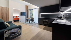 Una sala moderna con un sofá, una mesa de centro con libros, una zona de comedor con una mesa y sillas, y una cocina con encimera negra y electrodomésticos de acero inoxidable.
