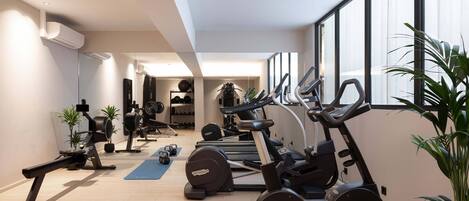 A modern gym with various exercise equipment, large windows, and indoor plants.