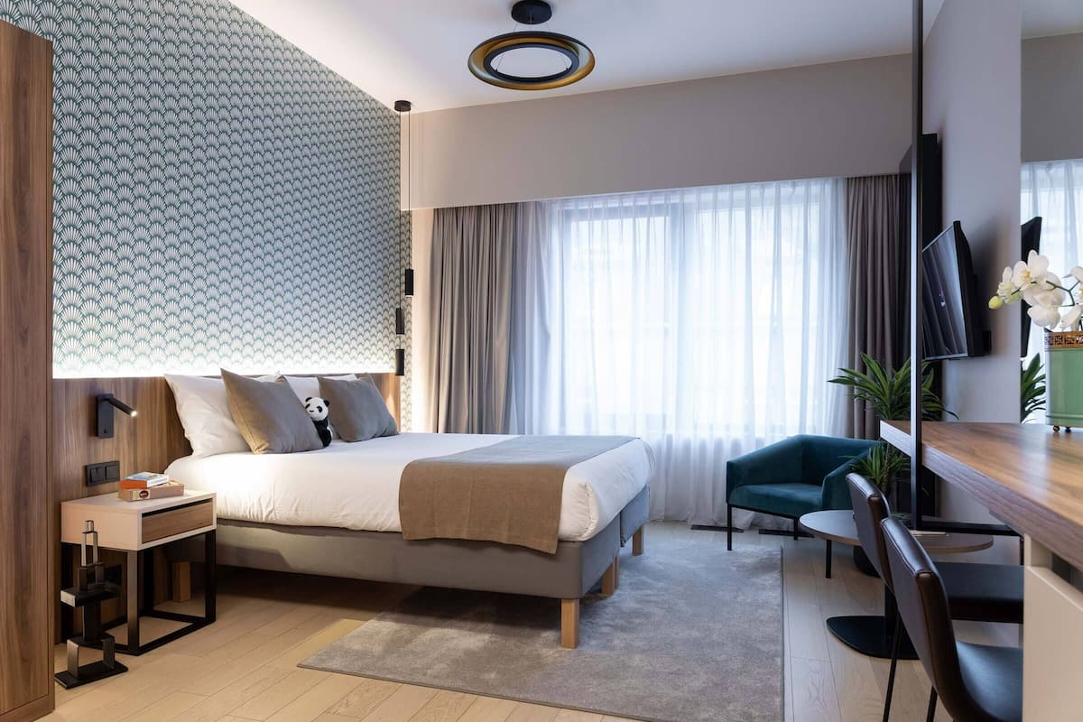 A modern hotel room with a bed, bedside table, lamp, TV, and a desk with a chair.