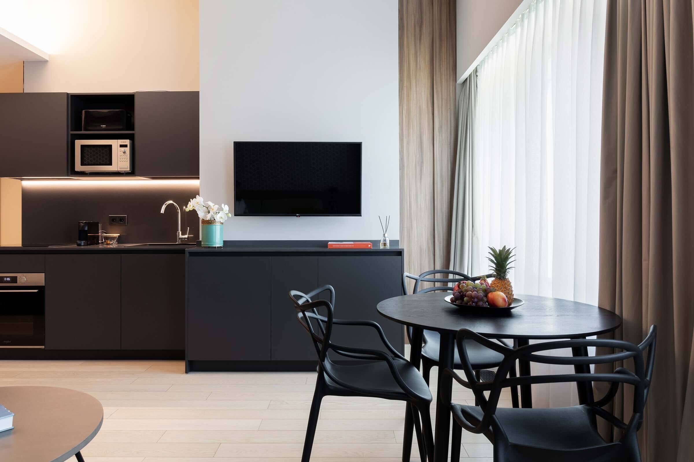 Ett modernt kök med en platt-TV, en mikrovågsugn och en kaffemaskin. Det finns en matplats med ett runt bord och stolar, och en fruktskål på bordet.