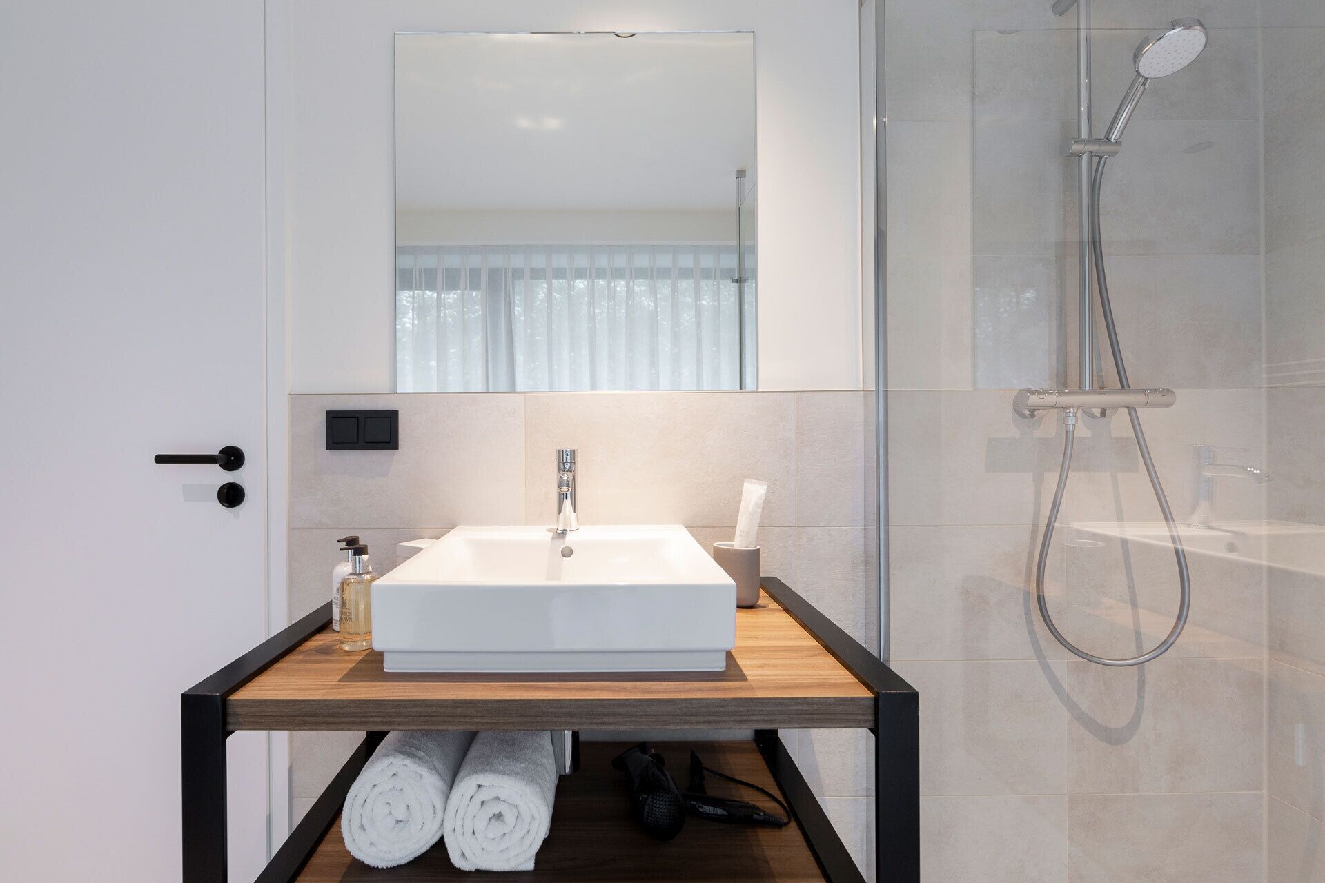 Un baño moderno con un lavamanos blanco, un espejo encima y una ducha a la derecha. Hay toallas y artículos de aseo en el estante del lavamanos.