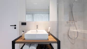 Un baño moderno con un lavamanos blanco, un espejo encima y una ducha a la derecha. Hay toallas y artículos de aseo en el estante del lavamanos.