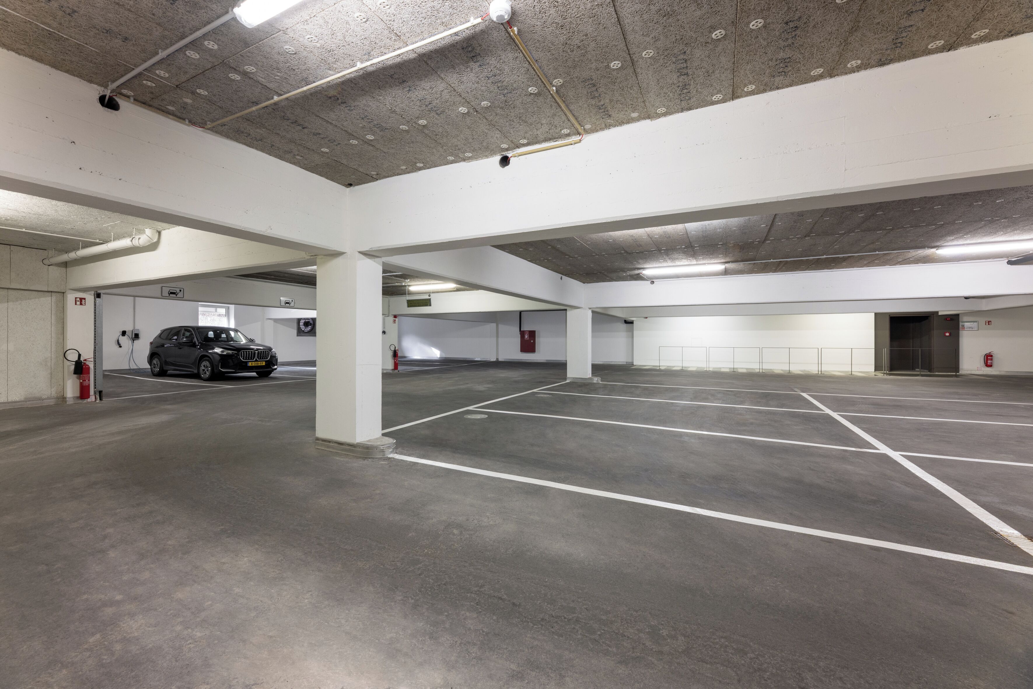 A multi-level parking garage with concrete floors and white walls, featuring several parking spaces and fire extinguishers.
