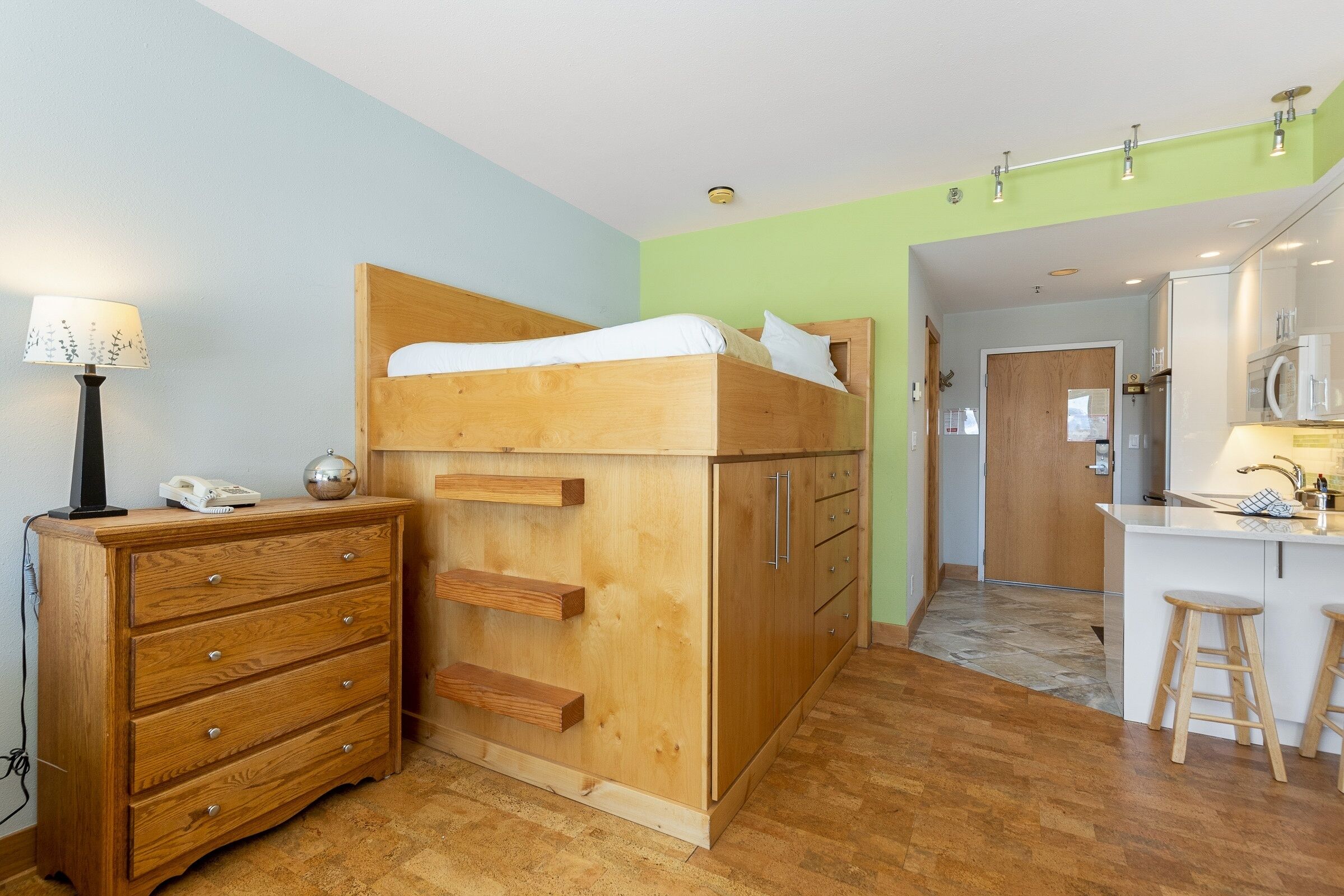 A bunk bed with a wooden headboard, a dresser with drawers, a kitchen area with a sink and stools, and a door leading to another room.