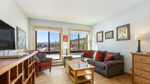 Uma sala de estar moderna com uma mesa de centro de madeira, um sofá cinzento e uma TV sobre um suporte de madeira. Há duas janelas com vista para as montanhas, e um armário de madeira com gavetas.