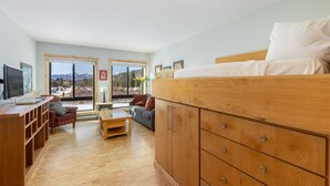 Quarto moderno com cama, móveis de madeira, uma escrivaninha e vista para montanhas através de grandes janelas.