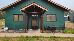 Uma cabana verde com um letreiro 'Cabin For Rent', uma porta da frente e um pequeno alpendre com mesa e cadeiras.