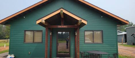 A green cabin with a 'Cabin For Rent' sign, a front door, and a small patio with a table and chairs.