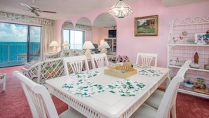 Una habitación con una mesa blanca, sillas, un ventilador de techo, una araña, un ventanal con vista al océano y una pared rosa.