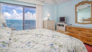 Un dormitorio con cama, cómoda, televisor, espejo y vista al océano.