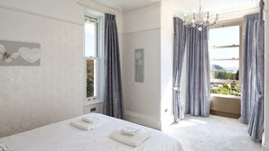 A bedroom with a large bed, two towels, a chandelier, and a view of the outdoors through a window with curtains. - Torbay Rise Seaside Villa with outstanding family facilities (Torquay)