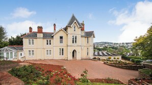 A large, two-story building with a symmetrical facade, a central entrance, and a landscaped garden with flower beds. - Torbay Rise Seaside Villa with outstanding family facilities (Torquay)