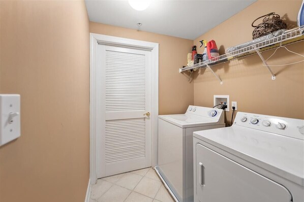 A laundry room with two washers and dryers, a white door, a shelf with cleaning supplies, and a light switch on the wall.