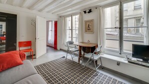 A room with a sofa, a patterned rug, a dining table with chairs, a TV, and large windows with curtains.