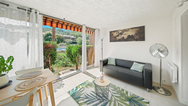 A modern living room with a sofa, a standing fan, and a view of greenery outside. - Charming T2 apartment near the sea - Residence with trees and swimming pool (Le Pradet)