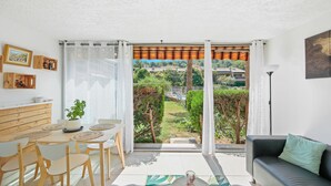 A modern living room with a dining area, a sofa, and a view of a garden and buildings. - Charming T2 apartment near the sea - Residence with trees and swimming pool (Le Pradet)
