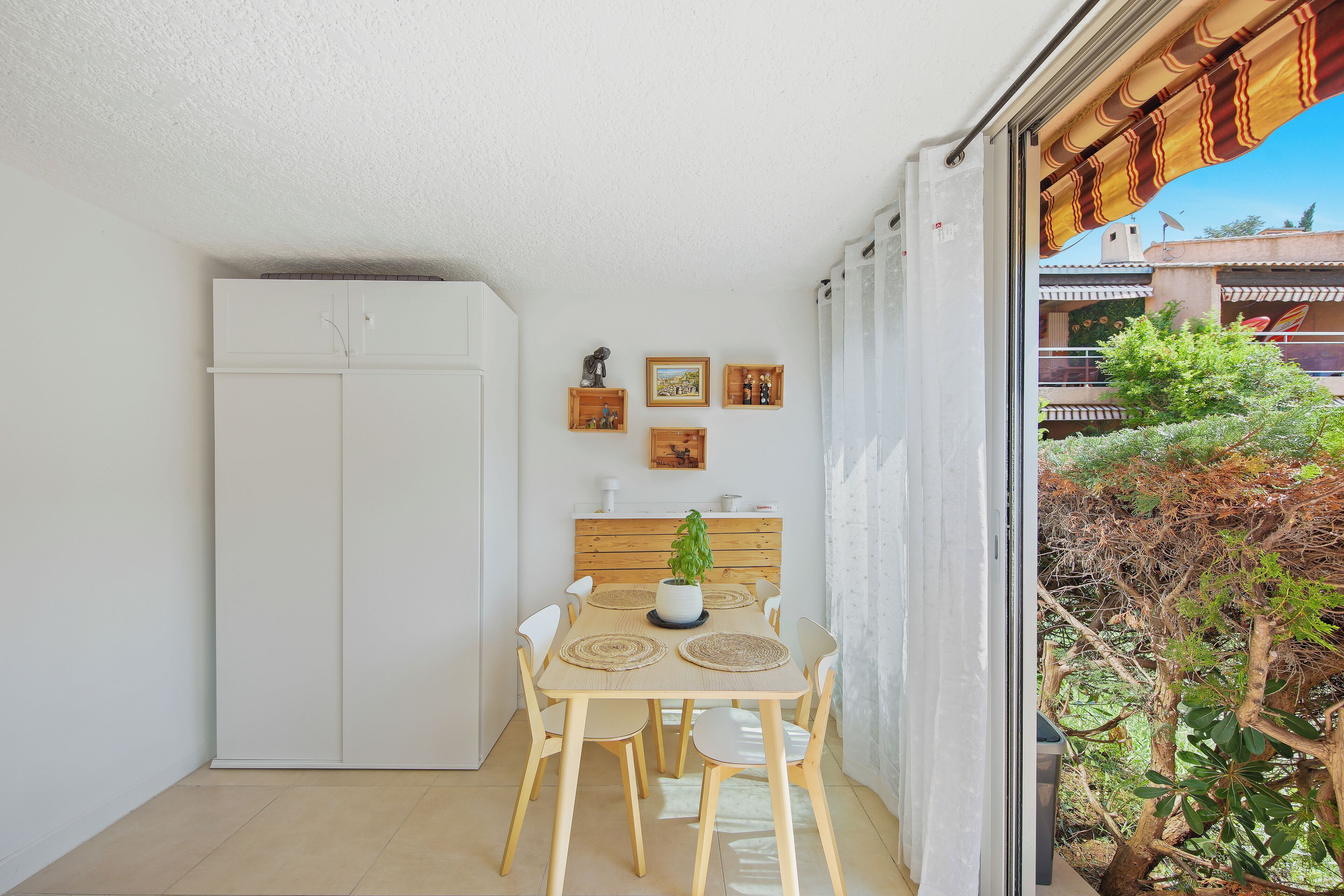 A small dining area with a table, chairs, and a potted plant. There is a white wardrobe and a window with a view of a tree and buildings.