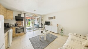 A modern kitchen with a dishwasher, oven, and coffee maker. There is a dining area with a table and chairs, and a living room with a sofa and a TV. - Charming T2 apartment near the sea - Residence with trees and swimming pool (Le Pradet)