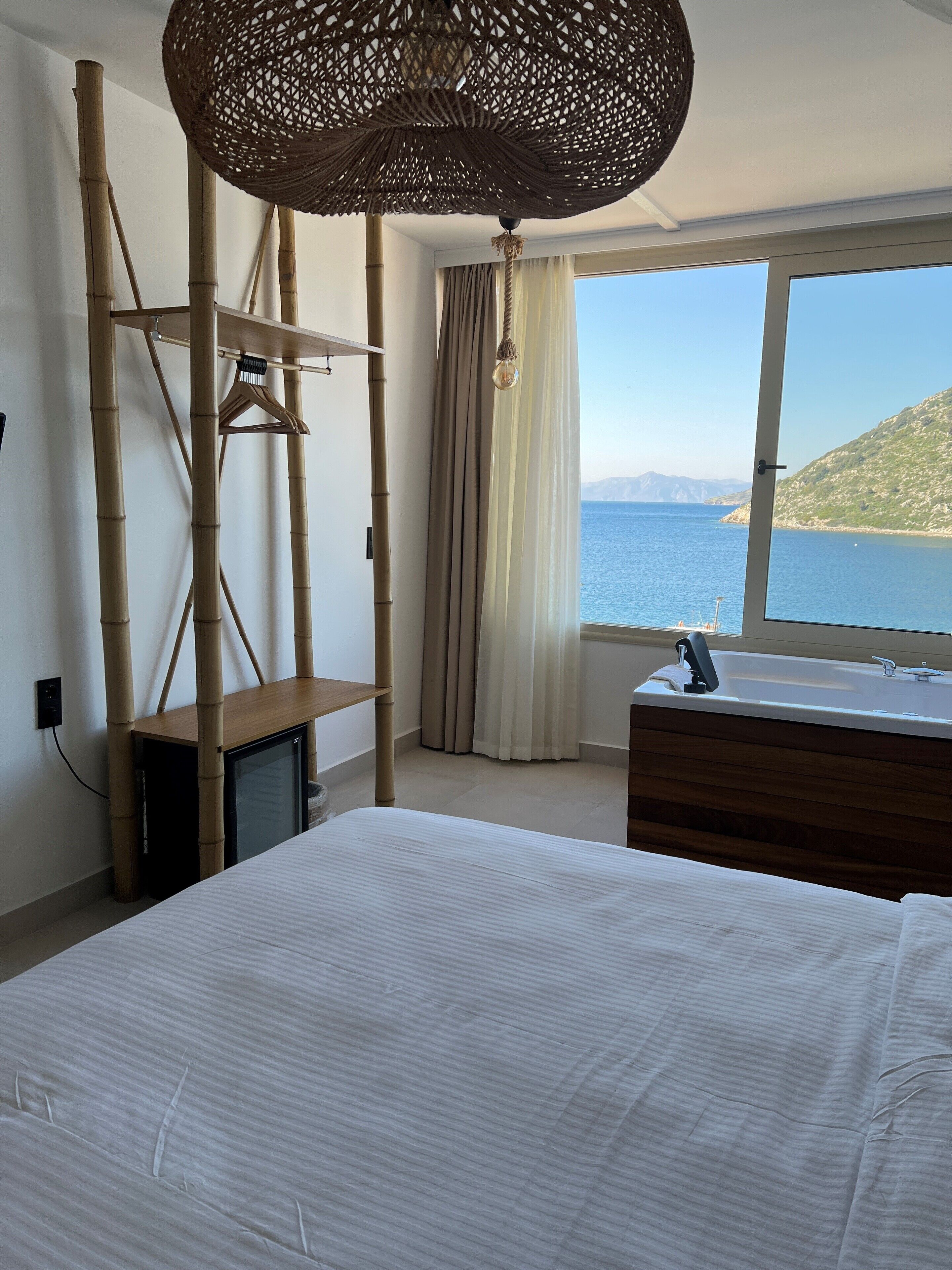A bedroom with a bed, a dresser, a TV, a large window with a view of the sea, and a hanging light fixture.