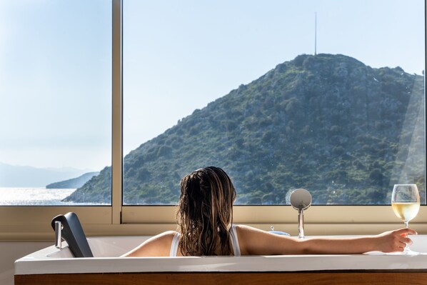 A person relaxing in a bathtub with a view of a mountainous landscape through a window. - Liman Hotel Gumusluk- Adult Only (Bodrum)