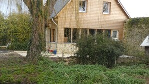 A wooden house with a pond in front, surrounded by greenery and a stone wall. - In the heart of D-Day" cottage (Barbeville)