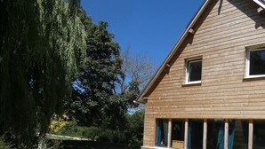 A wooden house with a stone foundation, a patio with outdoor furniture, and a well-maintained lawn. - In the heart of D-Day" cottage (Barbeville)