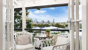 A balcony with white wicker chairs and a small table, overlooking a cityscape with buildings and greenery. - Family Pool Home | River Fire City Skyline Views (Balmoral)