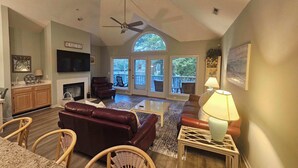 A spacious living room with a fireplace, leather sofas, a dining area with wicker chairs, and large windows offering a view of the outdoors.