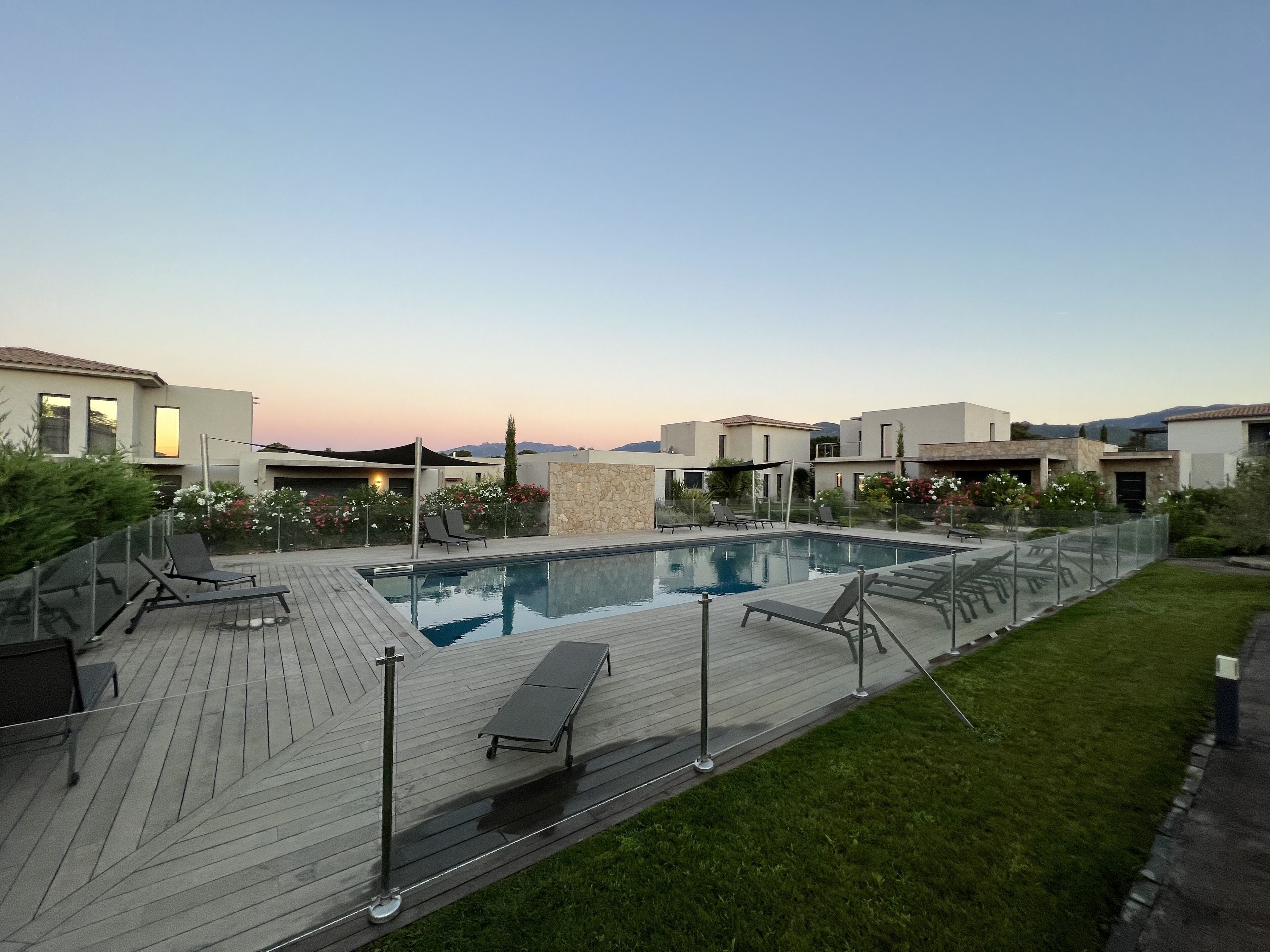 Een modern zwembad met ligbedden en een houten terras, omringd door verzorgde gazons en een heldere lucht.
