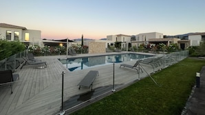 Ein moderner Poolbereich im Freien mit Liegestühlen und einer Holzterrasse, umgeben von gepflegten Rasenflächen und blauem Himmel.